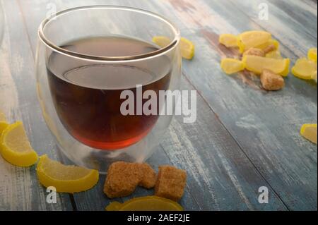 Marmalade lemon slices, pieces of brown sugar and a glass of black tea on a wooden background. Sweet dessert. Close up Stock Photo