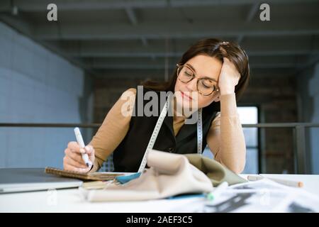 Young pensive female designer thinking over new fashion sketch in studio Stock Photo