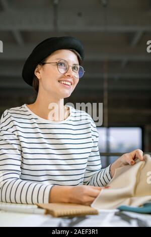Happy young female designer with piece of textile working by desk in studio Stock Photo