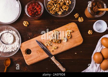 Overhead view chopped walnuts on cutting board with knife next to ingredients for baking brownie cake on brown wooden table. Stock Photo