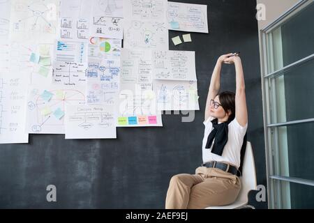 Young tired businesswoman stretching arms while sitting on chair by blackboard Stock Photo