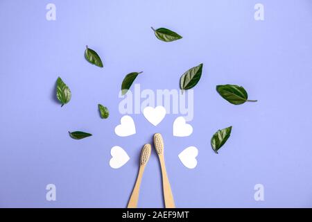Two wooden bamboo eco friendly toothbrushes, green leaf, white heart on purple background. Dental care, eco friendly, reuse and valentines day concept Stock Photo