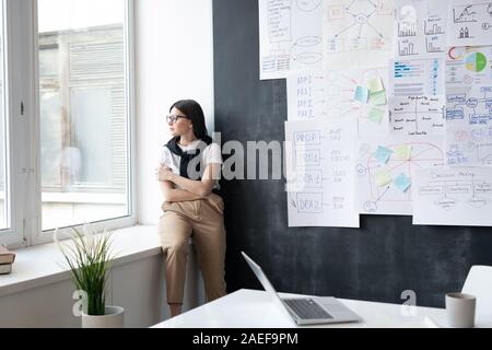 Young tired businesswoman in casualwear looking through window in office Stock Photo