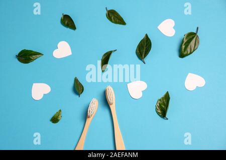 Two wooden bamboo eco friendly toothbrushes, green leaf, white heart on blue background. Dental care, eco friendly, reuse and valentines day concept. Stock Photo