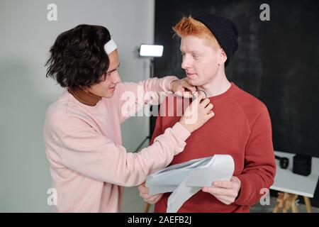 Casual guy putting microphone on sweatshirt of young male vlogger with paper Stock Photo