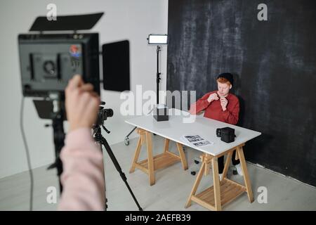 Young vlogger in casualwear sitting by desk against black background in studio Stock Photo