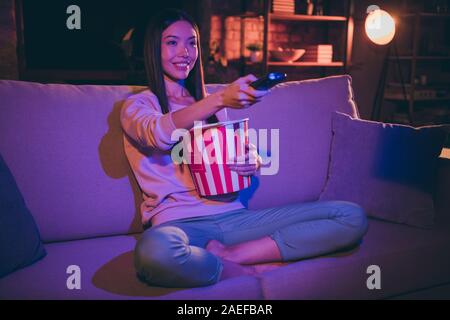 Photo of pretty lady domestic mood holding tv remote controller changing channels eating popcorn movie night sitting comfy couch casual outfit dark Stock Photo