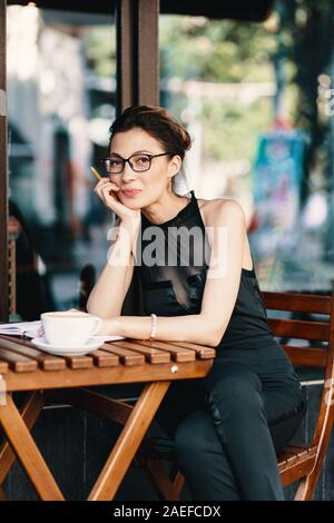 Young stylish woman with stylish glasses sitting at table in cafe with a big cup of coffee or latte, smiling studying, workplace, holding pencil Stock Photo