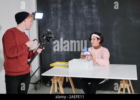 Young vlogger showing new footwear to his friend with video camera in front Stock Photo