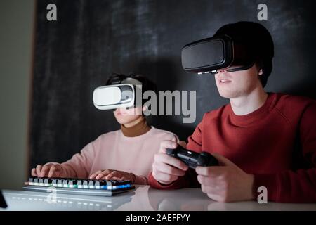 Two contemporary teenagers in vr goggles sitting in front of computer screen Stock Photo
