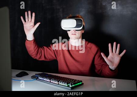 Young man in vr headsets sitting by desk in front of virtual computer screen Stock Photo