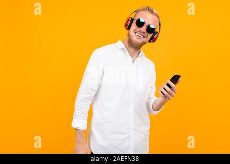 European guy in a white shirt and sunglasses listens to music in large headphones on a yellow background. Stock Photo