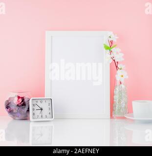 Blank white frame, flower, clock and cup of coffee or tea on a white table against the pink wall with copy space. Mock up. Stock Photo