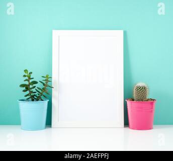 Blank white frame, plant cactus on a white table against the blue wall. Mockup with copy space. Stock Photo