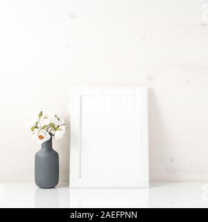 Blank white frame, flower in vaze on a white table against the white wall with copy space. Mock up. Stock Photo