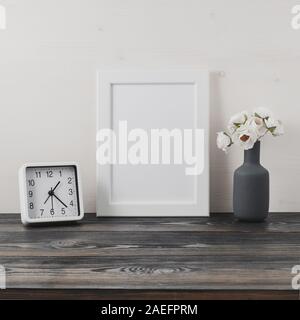 White frame, flower in vaze, clock on a dark gray wooden table against the white wall with copy space. Mock up. Stock Photo