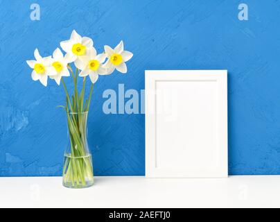 Bouquet of fresh flowers, daffodils with a glass vase on a white table, opposite the blue textured concrete wall. Empty space for text. Stock Photo