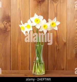 Bouquet of fresh flowers, daffodils in vase in the middle of wooden table, opposite brown wooden wall. Empty space for text. Stock Photo