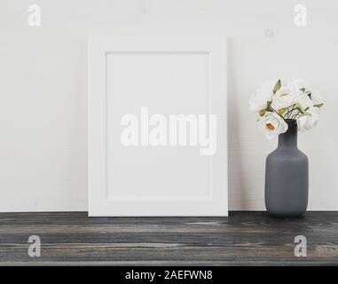 White frame, flower in the vase, clock on dark grey wooden table against the white wall with copy space. Mock up. Stock Photo