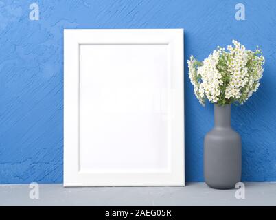 Blank white frame, flower in vaze on the gray table against dark blue concrete wall with copy space. Mock up. Stock Photo
