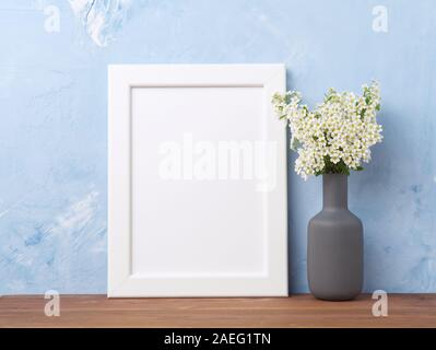 Blank white frame, flower in vaze on the brown wooden table against pastel blue concrete wall with copy space. Mock up. Stock Photo
