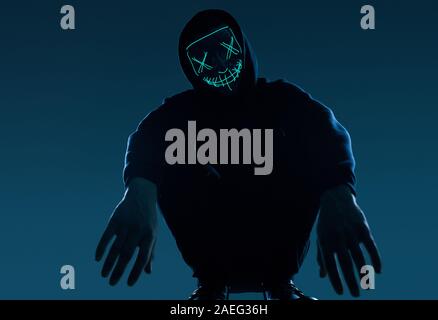 Portrait of an anonymous man in a black hoodie hiding his face behind a scary neon mask. Studio shot. Stock Photo