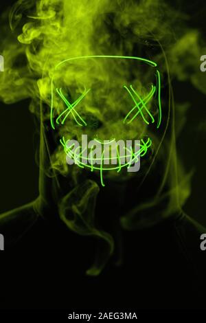 Close-up portrait of an anonymous man hiding his face behind a scary neon mask in a colored smoke. Studio shot Stock Photo