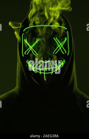 Close-up portrait of an anonymous man hiding his face behind a scary neon mask in a colored smoke. Studio shot Stock Photo