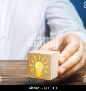 Businessman holds a wooden block with an image of a light bulb idea. Generation of innovative business ideas. Creative process. The accumulation of ex Stock Photo