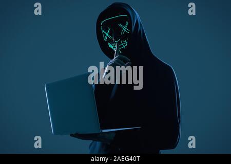 Portrait of an anonymous man in a black hoodie and neon mask hacking into a computer. Studio shot. Stock Photo