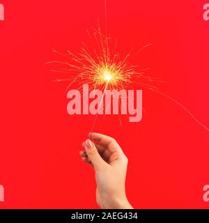 Female hand holds burning sparkler symbol of magic at Christmas on bright red background. Stock Photo