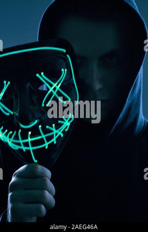 Portrait of an anonymous man in a black hoodie hiding his face behind a scary neon mask. Studio shot. Stock Photo