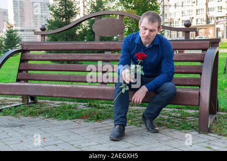 Pensive guy sitting on a bench with a rose in his hands in the park Stock Photo