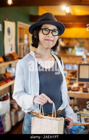 Japanese woman wearing hat and glasses standing in a leather shop, holding shopping bag, smiling at camera. Stock Photo