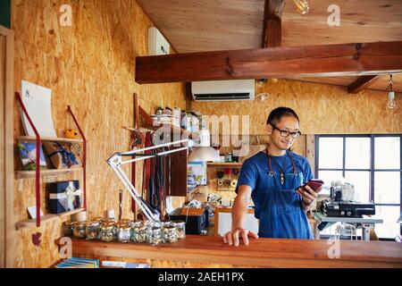 Japanese man wearing blue apron and glasses standing in a leather shop, using his mobile phone. Stock Photo
