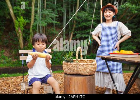 Japanese woman standing outdoors, wearing hat and apron and young boy sitting on chair, eating corn on the cob. Stock Photo