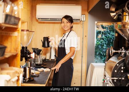 Japanese woman wearing apron standing in an Eco Cafe, preparing coffee, smiling at camera. Stock Photo