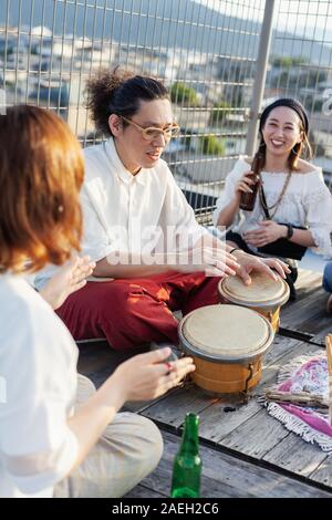 Two Japanese women and man sitting on a rooftop in an urban setting, playing drums. Stock Photo