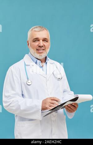 Portrait of medical professional in white coat standing and smiling at camera while writing in document isolated on blue background Stock Photo