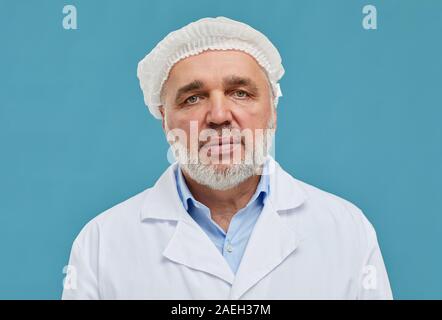 Portrait of mature surgeon in cap and in white coat looking at camera over blue background Stock Photo