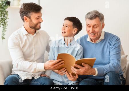 Boy Reading Book Sitting With Father And Grandfather On Sofa Stock Photo