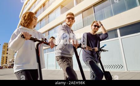 Three best friends young 20s -30s girl and guys spend time outdoors gathered together driving on electric scooter modern land vehicle Stock Photo