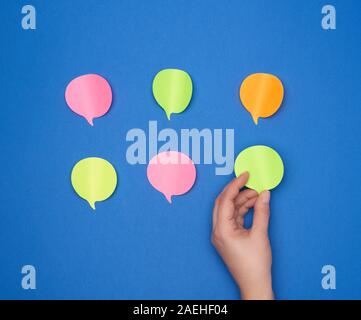 empty paper multi-colored stickers in the shape of a cloud and a female hand glues one piece of paper on a blue background, dialogue concept Stock Photo