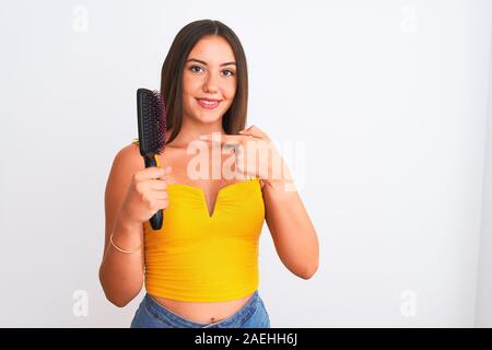 Young beautiful girl holding hair comb standing over isolated white background with surprise face pointing finger to himself Stock Photo