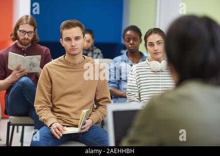 Young man sitting on chair and listening to his teacher with other students in the class Stock Photo