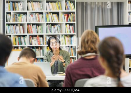 Asian young teacher in eyeglasses sitting at desk and explaining the new material to the group of students in the library Stock Photo