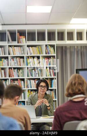 Young man sitting at the table and talking to the group of students at the university library Stock Photo