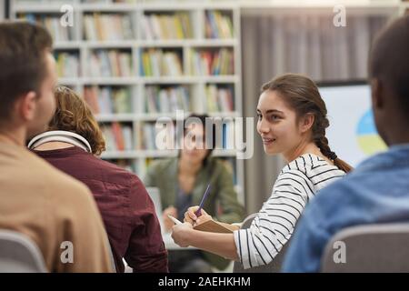 Young woman making notes in her notebook and discussing it with her colleagues during business presentation Stock Photo
