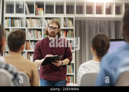 Young teacher in eyeglasses standing and talking to the students in the university library Stock Photo
