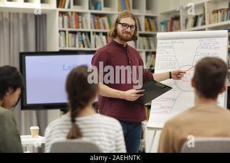 Young man in eyeglasses standing near the whiteboard and teaching the young people during presentation at library Stock Photo